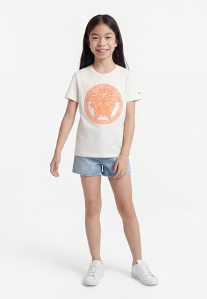 Fată zâmbitoare cu păr lung și întunecat, purtând un tricou alb cu imprimeu portocaliu cu capul Medusei, pantaloni scurți de blugi și adidași albi, stând pe un fundal alb.