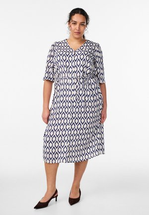 Žena stojaca v bielych šatách po kolená s námornícky modrým diamantovým vzorom, zapínaných na gombíky vpredu, s polovičnými rukávmi a s čiernymi nízkymi opätkami.