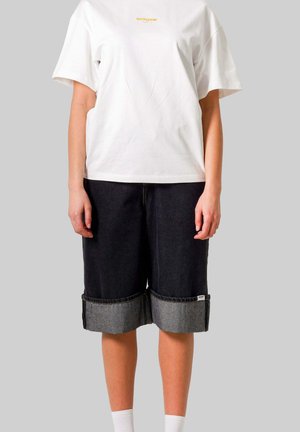 Persoon draagt een wit shirt met korte mouwen met kleine gele tekst en donkere opgerolde denimshorts tegen een effen grijze achtergrond.