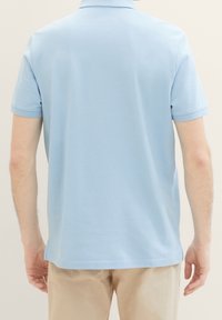 Jasnoniebieska koszulka polo z krótkim rękawem, wykonana z gładkiej tkaniny, z klasycznym kołnierzykiem i subtelną fakturą. Bez widocznych logo ani wzorów.