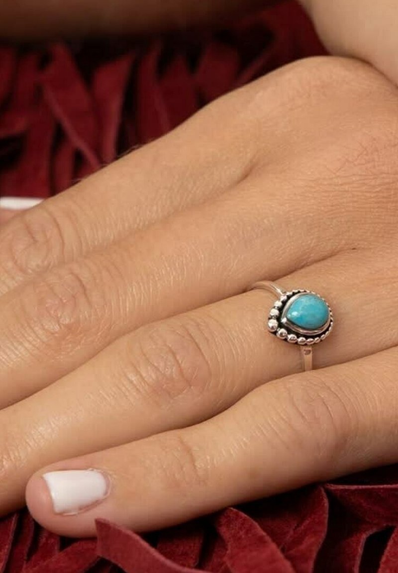 Main à peau claire portant une bague en argent ornée d'une pierre turquoise en forme de goutte, reposant sur un tissu rouge foncé texturé.