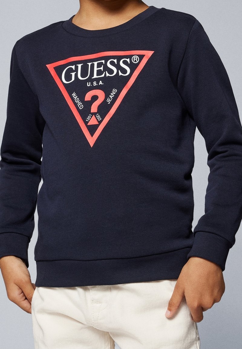 Enfant portant un sweat-shirt bleu marine avec un logo triangulaire rouge et le texte blanc « GUESS U.S.A. », assorti à un pantalon de couleur claire.