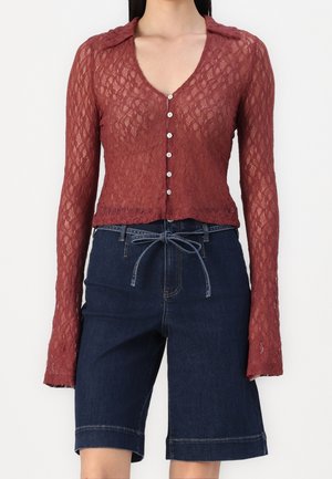 Mujer viste una blusa de encaje color óxido semitransparente con botones y mangas largas, y shorts vaqueros azul oscuro hasta la rodilla con un lazo en la cintura.