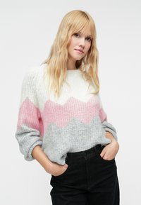 Adīts džemperis ar zigzaga rakstu baltā, rozā un pelēkā krāsā. Audums ir mīksts un teksturēts, ar uzvērtiem piedurknēm un brīvu piegriezumu.