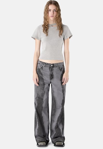 Jeune femme aux cheveux bouclés portant un crop top gris clair à manches courtes et un jean large noir et gris délavé avec des chaussures à plateforme.