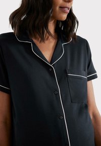 Czarna koszula z krótkim rękawem, zapinana na guziki, z białym obszyciem przy kołnierzyku i kieszeni, wykonana z miękkiego materiału, z jednym detalem kieszeni na piersi.