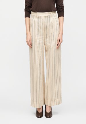 Kvinde iført cremefarvede, højtaljede bukser med vide ben og tynde lodrette striber, kombineret med en brun langærmet top og brune sko.