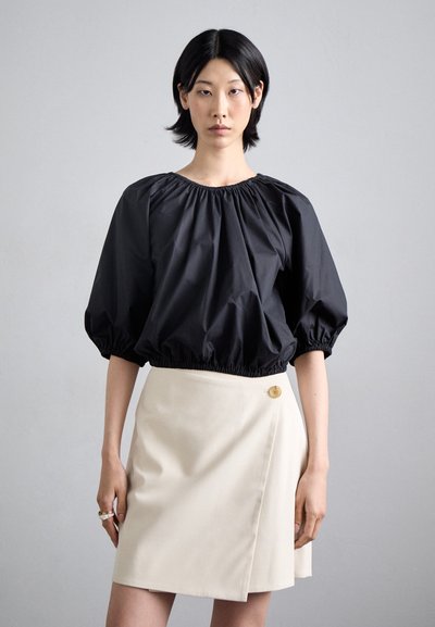 Blouse noire cropped avec des manches bouffantes et un ourlet élastique, associée à une jupe enroulée beige avec un détail de bouton proéminent.