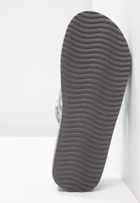 Semelle de chaussure en caoutchouc noir avec une texture ondulée, dotée d'une tige grise et duveteuse. La semelle s'étend légèrement vers l'extérieur au talon, créant une forme arrondie.