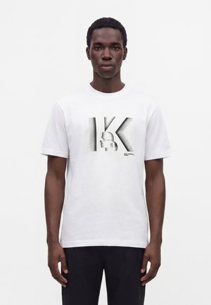 Άνδρας με λευκό μπλουζάκι με μεγάλο μαύρο "K" που ξεθωριάζει και λογότυπο σιλουέτας, στέκεται μπροστά σε απλό λευκό φόντο.