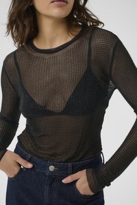 Vrouw in een zwart doorschijnend long-sleeve shirt met kleine zwarte studs over een zwarte bralette en blauwe jeans, met haar handen in de zakken.