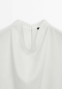 Blouse blanche avec un col rond et deux petits boutons au dos pour la fermeture, en tissu léger.