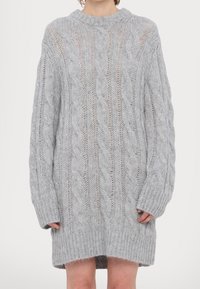 Pull gris clair surdimensionné en maille torsadée avec manches longues, poignets et ourlet côtelés, porté comme une robe par une personne debout.