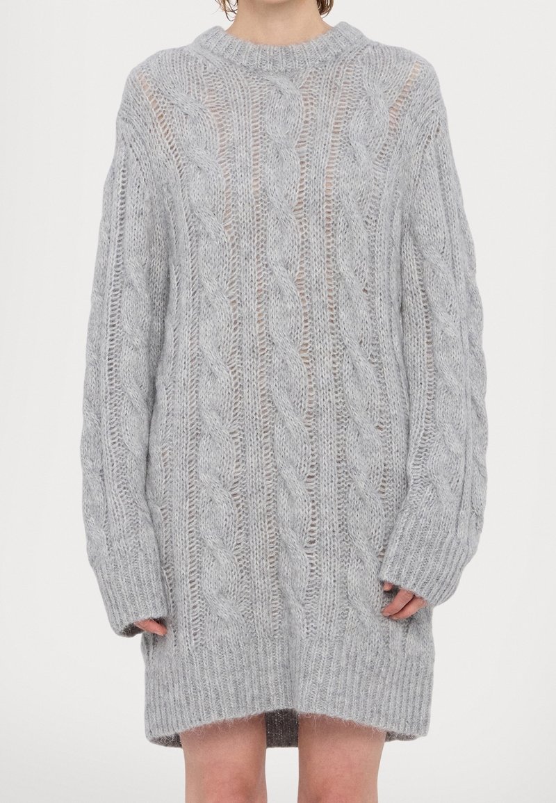 Pull gris clair surdimensionné en maille torsadée avec manches longues, poignets et ourlet côtelés, porté comme une robe par une personne debout.