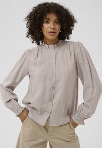 Lys grå langærmet bluse med rynket krave, knap foran og samlede ærmer. Stoffet ser blødt ud med en glat tekstur.