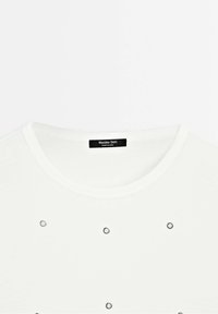 T-shirt blanc à col rond avec cinq petits œillets métalliques argentés également espacés sur la poitrine et une étiquette noire à l'intérieur du col.