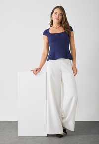 Femme portant un haut ajusté bleu marine et un pantalon large blanc, se tenant à côté d'un grand panneau rectangulaire blanc sur un sol gris.