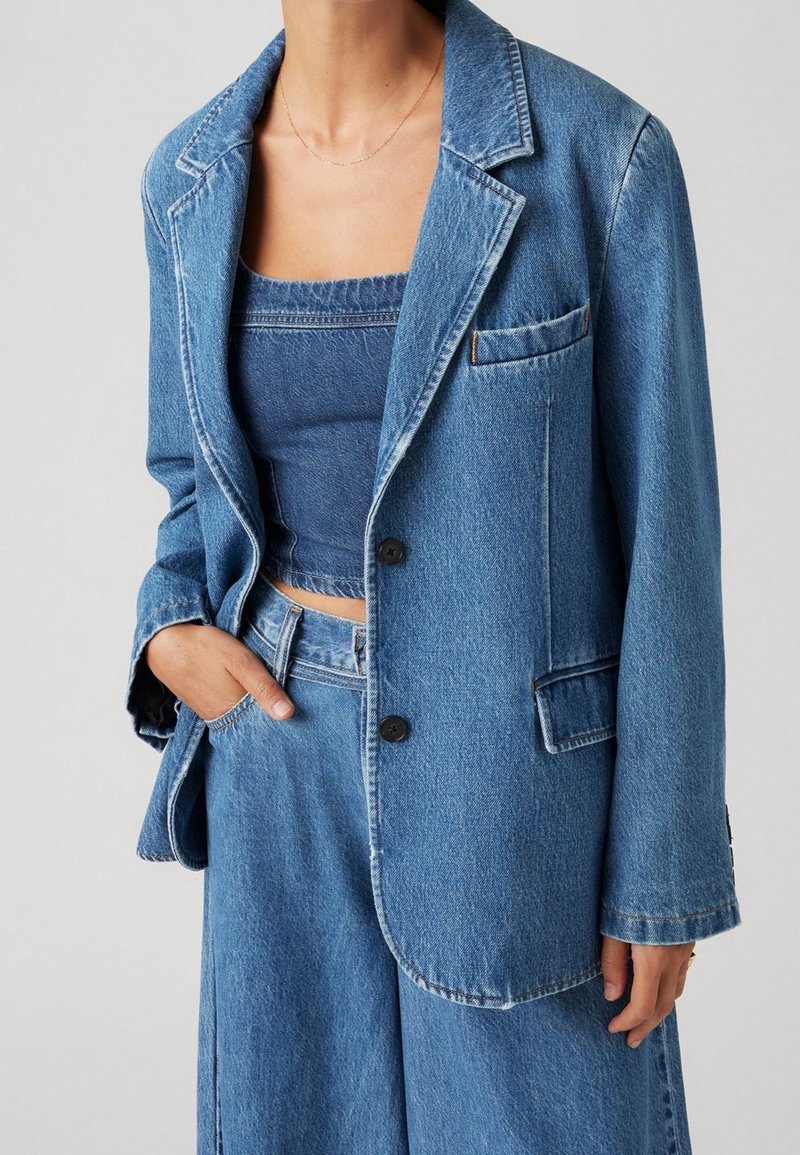 Femme portant un blazer en denim bleu, un jean large assorti et un crop top en denim, une main dans la poche.