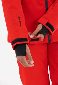 Rode waterdichte jas met een zwarte binnenmanchet, voorzien van klittenbandaccenten en een label met de tekst "W-PRO 15.000" op de mouw.