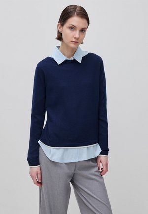 Donna con capelli corti castani che indossa un maglione blu navy sopra una camicia celeste con colletto e pantaloni grigi, in piedi contro uno sfondo semplice.