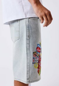 Persona con pantalones cortos de mezclilla claros con parches bordados de colores y una camisa blanca, visible desde la cintura hasta la rodilla sobre un fondo liso.