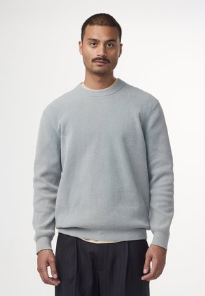 Felpa grigia chiara con texture, colletto a girocollo, polsini e orlo a coste, indossata sopra una camicia color crema, abbinata a pantaloni scuri a righe.