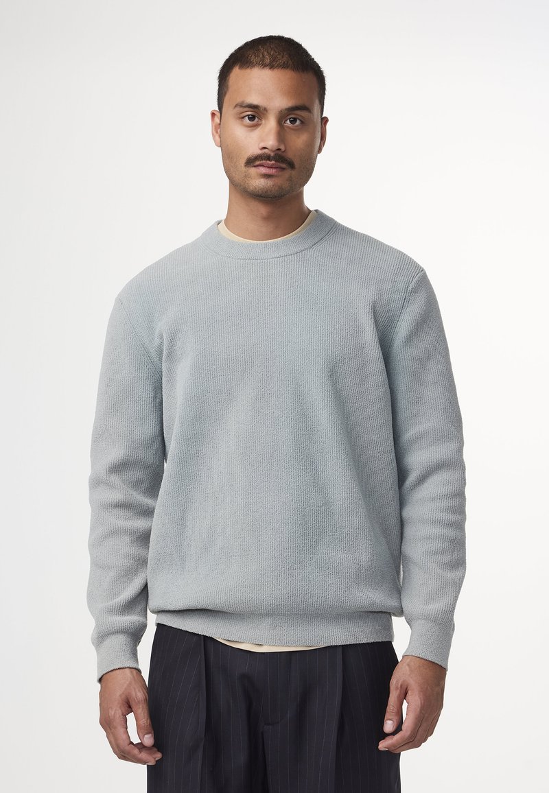Hellgrauer Sweatshirt mit Struktur, Rundhalsausschnitt, gerippten Bündchen und Saum, getragen über einem cremefarbenen Oberteil, kombiniert mit dunkel gestreiften Hosen.