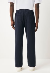 Asmuo dėvi tamsiai mėlynas kelnes su galinėmis kišenėmis ir baltą marškinius, stovi priešais paprastą šviesų foną.