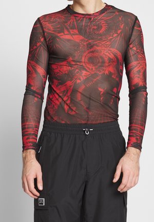 Homme portant un haut noir transparent à manches longues avec des motifs abstraits rouges et un pantalon cargo noir à taille cordon.