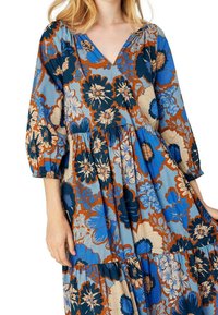 Robe maxi fleurie en bleu, marron et crème. Comprend un col en V, des manches bouffantes et une jupe à volants avec des motifs floraux variés.