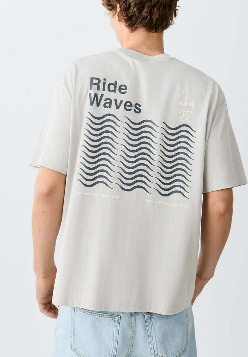 Personne portant un t-shirt gris clair avec "Ride Waves" et des motifs en forme de vagues imprimés dans le dos, associé à un jean bleu clair.