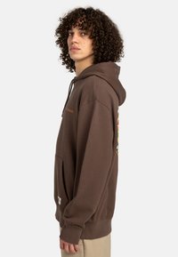Sweatshirt à capuche marron en matériau doux, doté d'une poche kangourou et de détails de design graphique au dos.