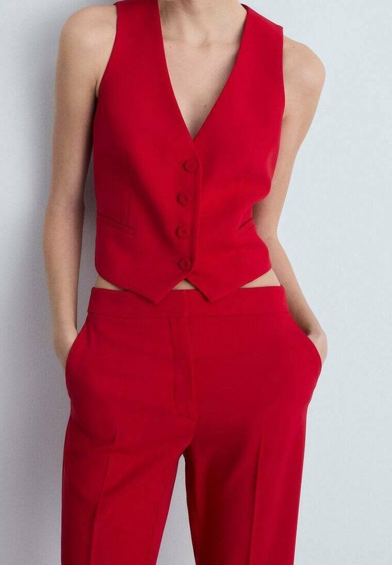 Czerwona kamizelka bez rękawów z guzikami, zestawiona z czerwonymi spodniami z wysokim stanem, ręce w kieszeniach, na jednolitym jasnym tle.