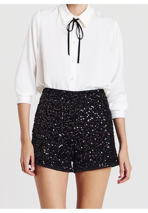 Femme portant un chemisier blanc boutonné avec un col orné de perles et un nœud noir, associé à un short noir à sequins taille haute.