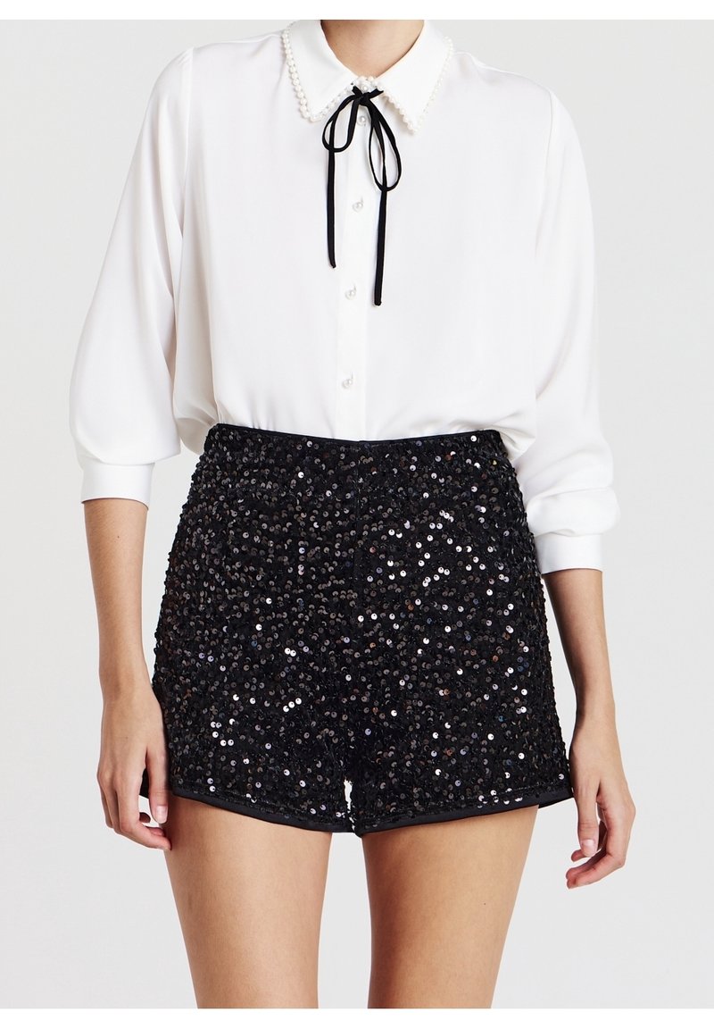 Femme portant un chemisier blanc boutonné avec un col orné de perles et un nœud noir, associé à un short noir à sequins taille haute.