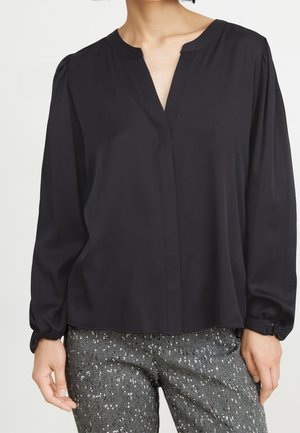 Frau trägt eine lockere schwarze Langarmbluse mit einem ausgeschnittenen Ausschnitt und graue gemusterte Hose mit weißen Sprenkeln.