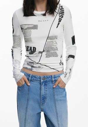 Femme portant une chemise blanche à manches longues avec un texte noir et des impressions graphiques, associée à un jean bleu taille haute, les mains dans les poches.