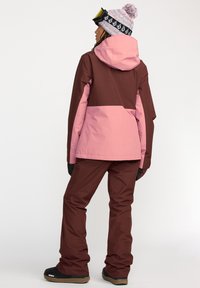 Chaqueta de esquí en marrón y rosa con capucha, con mangas largas y pantalones a juego. El conjunto se completa con guantes y botas negras.