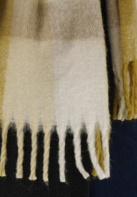 Uldent ternet tørklæde i gul, creme og beige; har frynser langs kanterne og en blød, tekstureret overflade. Egnet til varme i køligere vejr.