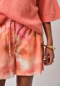 Korte mouwen, zachtroze trui met een losse pasvorm; gecombineerd met oranje tie-dye shorts met een trekkoord en metallic accenten op de koorden.