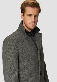 Grau gewebter Mantel mit strukturierter Oberfläche, mit Reverskragen und zwei Knöpfen, über einer schwarzen Zip-Jacke getragen.