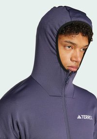 Veste à capuche zippée en tissu bleu foncé avec une texture lisse. Comprend une capuche ajustée et un logo "TERREX" blanc sur la poitrine.