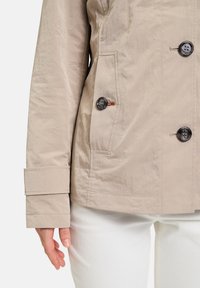 Beige Windbreakerjacke mit strukturierter Oberfläche, ausgestattet mit schwarzen Knöpfen und seitlichen Taschen. Mit Bündchenärmel und klaren Linien.