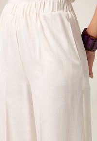 Pantaloni largi, de culoare bej deschis, confecționați dintr-un material moale, cu o talie elastică și buzunare laterale. Textură notabilă și efect de cădere.