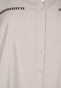 Camicia button-up in tessuto beige chiaro con finitura testurizzata, caratterizzata da due tasche sul petto e una fila di borchie argentate decorative lungo le spalle.