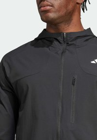Chaqueta con capucha negra de acabado texturizado, que cuenta con una cremallera frontal completa, un bolsillo adicional con cremallera y un acento de logo en el pecho.
