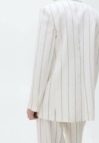 Personne portant un costume couleur crème à fines rayures verticales noires, vue de dos, avec un blazer et un pantalon assorti visibles.