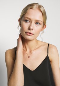 Collier en or rose avec un pendentif en forme de cœur orné de pierres précieuses roses, porté avec un camisole noir. Texture lisse et chaîne délicate.