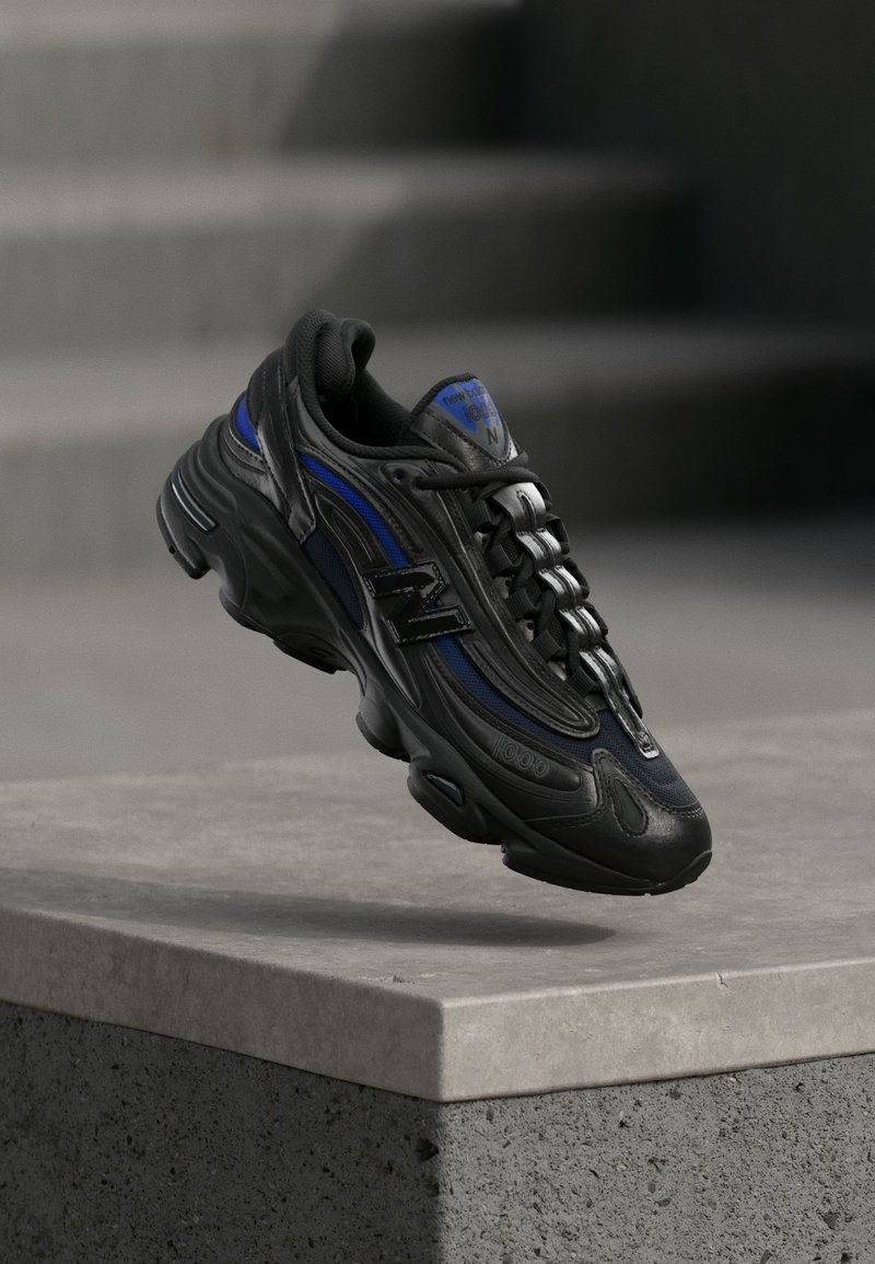 Μαύρο και μπλε παπούτσι New Balance που αιωρείται πάνω από μια τσιμεντένια άκρη με σκάλες στο θολό παρασκήνιο.