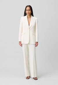 Blazer blanco a medida con solapas de muesca y bolsillos de solapa, combinado con pantalones anchos a juego. Textura suave, diseño minimalista.